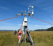 Two researchers stand under an instrument tower and reach up to a box on the tower.