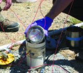 A canister with attached wires held on the ground by a gloved hand.