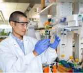 A research standing in a laboratory holds up a sample with gloved hands.