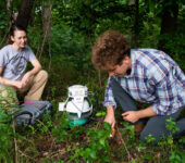 Two people with research instruments kneel in a forest floor, surrounded by vines and trees.