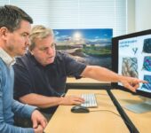 Two researchers sit in front of a computer and point at maps on the screen.